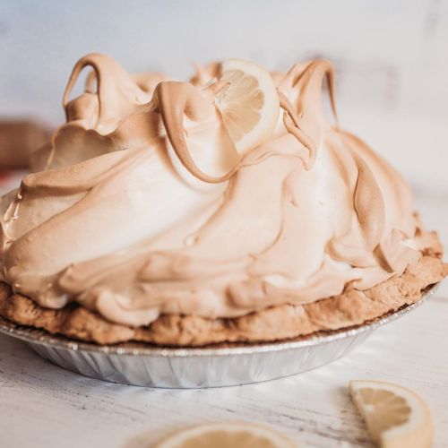 A lemon meringue pie with a golden, fluffy meringue topping and a slice of lemon on top, set on a white surface.
