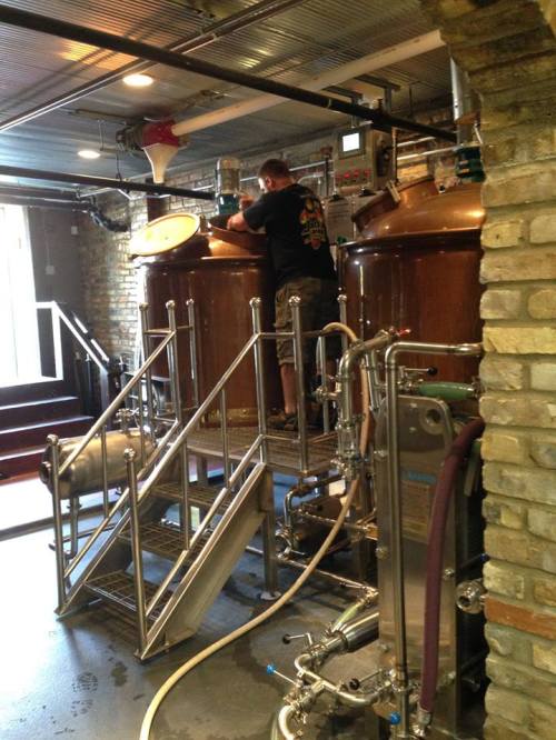 A person works on brewing equipment in a brewery, surrounded by large copper tanks and stainless steel machinery.