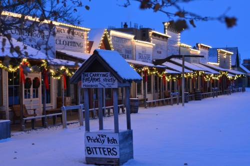A snowy street scene with festive lights and decorations on wooden buildings, creating a cozy winter atmosphere.
