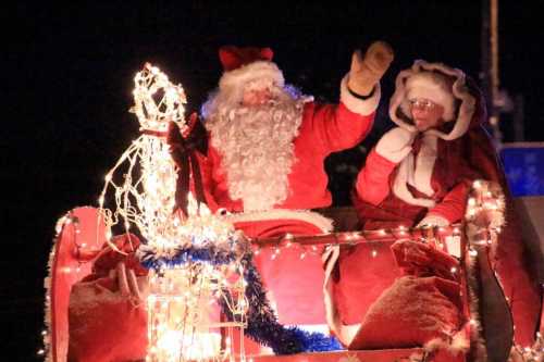 Santa Claus and Mrs. Claus wave from a decorated sleigh adorned with lights during a festive nighttime event.