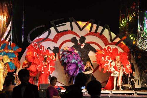 A vibrant stage with performers in colorful costumes and feathered headdresses, celebrating a Gemini-themed event.
