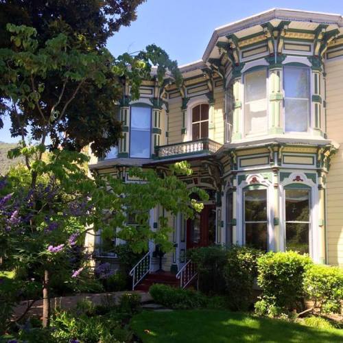 A colorful Victorian-style house surrounded by lush greenery and flowers, featuring intricate architectural details.