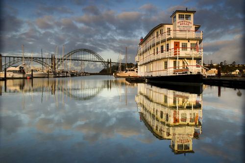 A historic building reflects in calm water, with a bridge and boats in the background under a cloudy sky.