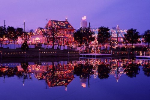 A beautifully lit house and trees reflect in a calm lake at dusk, adorned with colorful holiday lights.