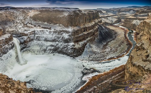 A panoramic view of a frozen waterfall surrounded by rocky cliffs and a winding river in a winter landscape.