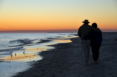 A couple walks along a beach at sunset, with gentle waves and a colorful sky in the background.