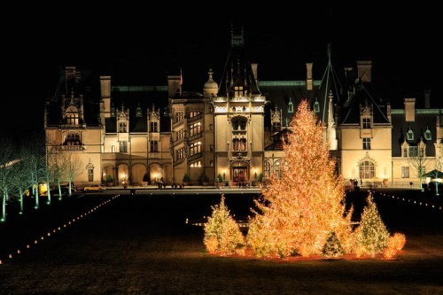 A grand mansion illuminated at night, surrounded by glowing Christmas trees and festive lights.