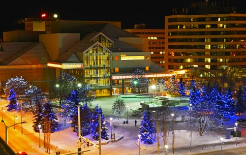 A snowy cityscape at night featuring a building with colorful lights and illuminated trees in a park.