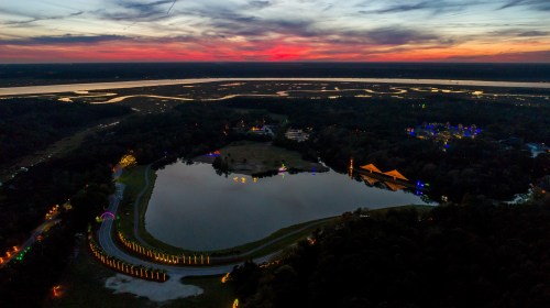 Aerial view of a serene lake at sunset, surrounded by trees and illuminated decorations, with winding waterways in the background.