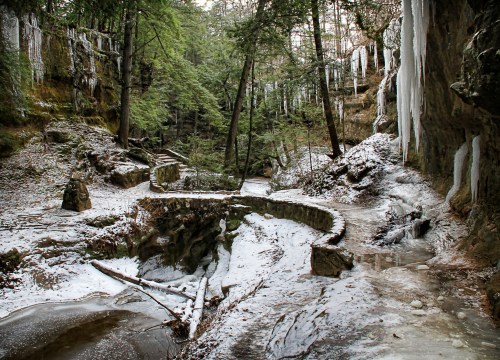 A serene winter landscape featuring icy rocks, frozen waterfalls, and snow-covered paths in a forested area.