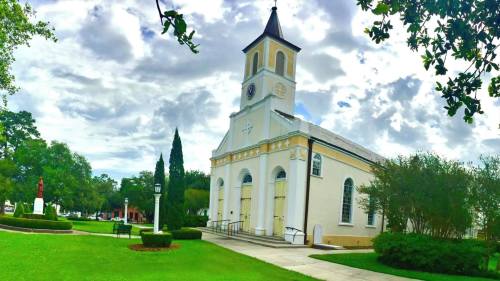 A historic church with a tall steeple, surrounded by greenery and a cloudy sky.