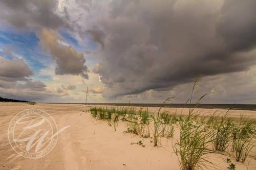 A sandy beach with sparse grass, under a dramatic sky filled with dark clouds and distant rain.