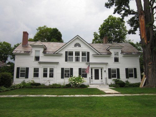 A large white house with black shutters, an American flag, and a well-maintained lawn under a cloudy sky.