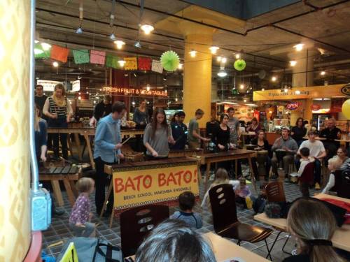 A lively performance with musicians playing marimbas, while an audience, including children, watches in a vibrant market setting.