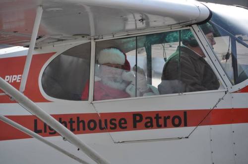 Santa Claus sits in the cockpit of a small plane, wearing a red suit and hat, with "Lighthouse Patrol" on the side.