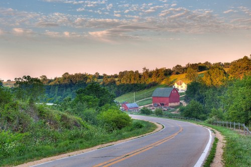 A winding road leads through lush greenery, with red barns and a yellow farmhouse in the background under a clear sky.