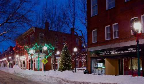 A snowy street at dusk, adorned with festive lights and decorations on buildings and a Christmas tree.