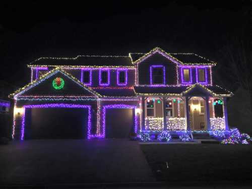 A house decorated with colorful Christmas lights, featuring purple and white strands, wreaths, and illuminated landscaping.