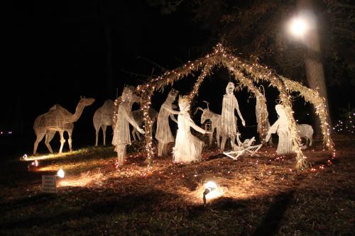A festive outdoor nativity scene with illuminated figures, camels, and twinkling lights under a night sky.