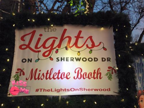Sign for "The Lights on Sherwood Dr" featuring a Mistletoe Booth, decorated with lights and greenery.