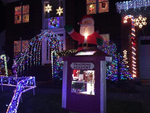 A festive house decorated with colorful lights, featuring a large Santa and a sign for a "Training Academy."
