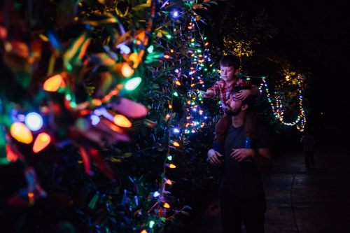 A man carries a boy on his shoulders, both admiring colorful holiday lights in a dark setting.