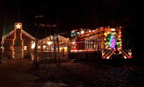 A brightly lit train and festive buildings adorned with colorful lights at night, surrounded by trees.