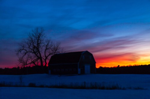 Silhouette of a barn and tree against a vibrant sunset sky with shades of blue, purple, and orange.