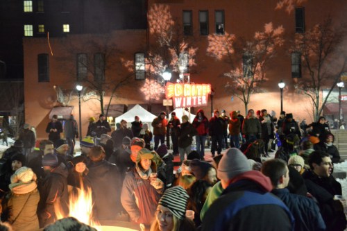 A lively winter festival scene with a crowd gathered around a fire, enjoying the festivities under twinkling lights.