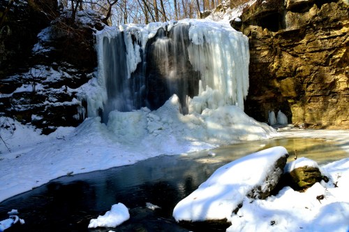 A frozen waterfall surrounded by snow-covered rocks and trees, with ice formations and a partially frozen pool below.