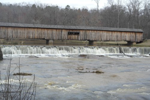 A wooden covered bridge spans a river with a waterfall, surrounded by bare trees and a cloudy sky.