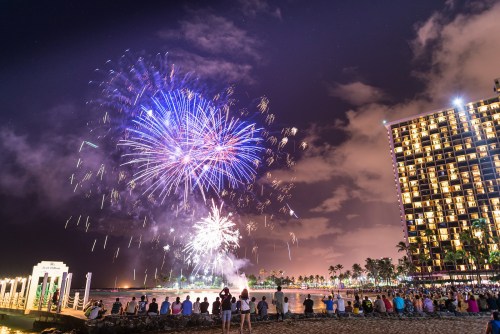 A vibrant fireworks display lights up the night sky over a beach, with spectators watching from the shore.