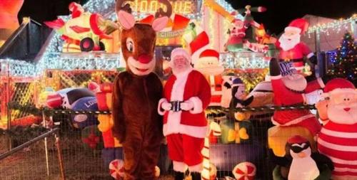 A person in a reindeer costume stands next to Santa Claus in front of a festive, brightly lit holiday display.