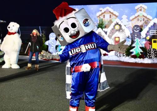 A cheerful mascot in a blue costume with a Santa hat poses in front of a festive winter backdrop with trees and decorations.