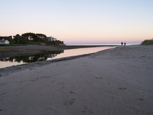 A serene beach at dusk, with two people walking along the shore and a calm river reflecting the pastel sky.