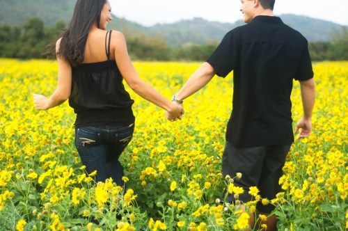 A couple walks hand in hand through a vibrant field of yellow flowers, enjoying a sunny day together.