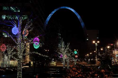 The Gateway Arch illuminated at night, surrounded by colorful holiday lights on trees and buildings.