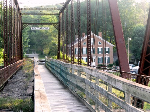 A rustic bridge leads to a brick house, surrounded by lush greenery and a calm waterway in the background.
