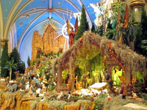 A vibrant nativity scene with figures, animals, and a backdrop of trees and mountains, set in a decorated church interior.