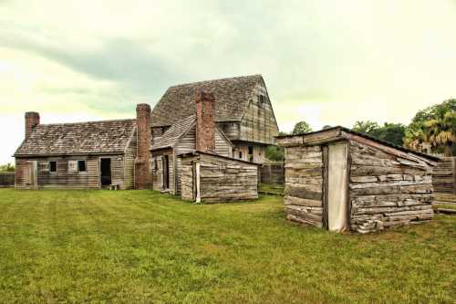 Historic wooden buildings with a grassy area, featuring a large house and smaller outbuildings under a cloudy sky.