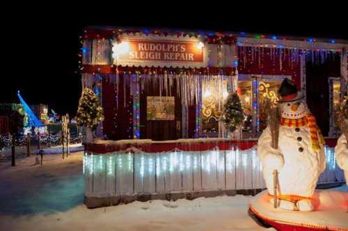 A festive scene featuring a sleigh repair shop decorated with lights and a snowman in a snowy landscape.