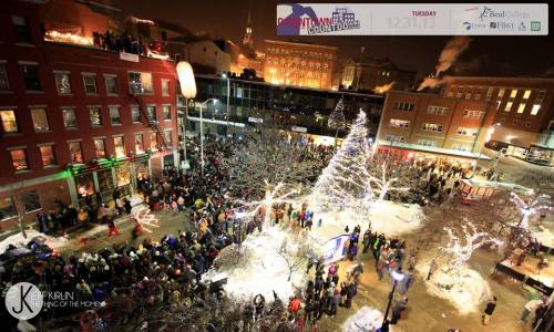 A festive crowd gathers in a snowy plaza, illuminated by holiday lights and a large Christmas tree.