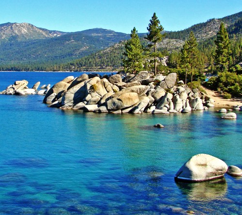A serene lake scene with clear blue water, rocky shores, and pine trees against a backdrop of mountains.