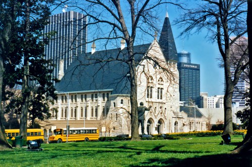 Historic building surrounded by trees, with modern skyscrapers and yellow school buses in the background.