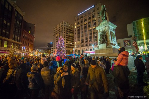 A large crowd gathers around a Christmas tree and monument at night, with festive lights and buildings in the background.