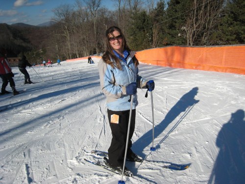 A woman smiles while skiing on a snowy slope, wearing a blue jacket and gloves, with trees and skiers in the background.