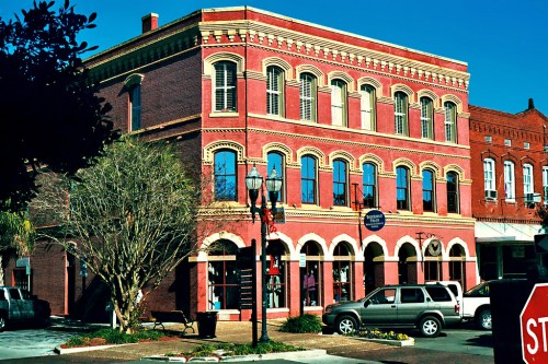 Historic red brick building with arched windows, surrounded by trees and parked cars, under a clear blue sky.