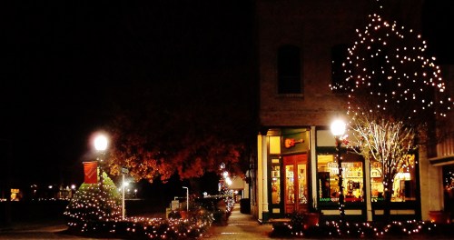 A festive street scene at night, adorned with twinkling holiday lights on trees and buildings.