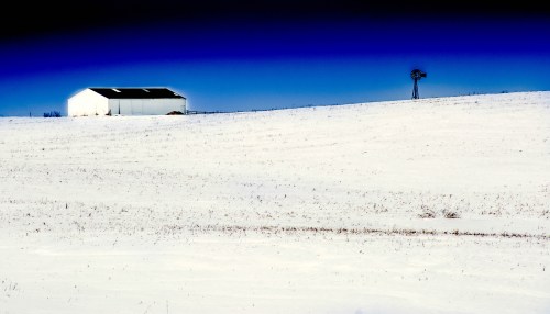 A white barn stands on a snowy landscape under a deep blue sky, with a lone windmill in the distance.