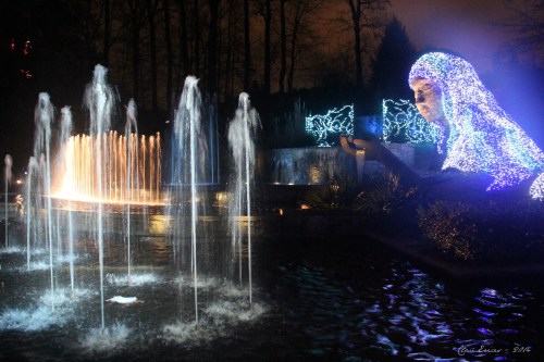 A serene nighttime scene featuring illuminated fountains and a large, glowing statue surrounded by trees.